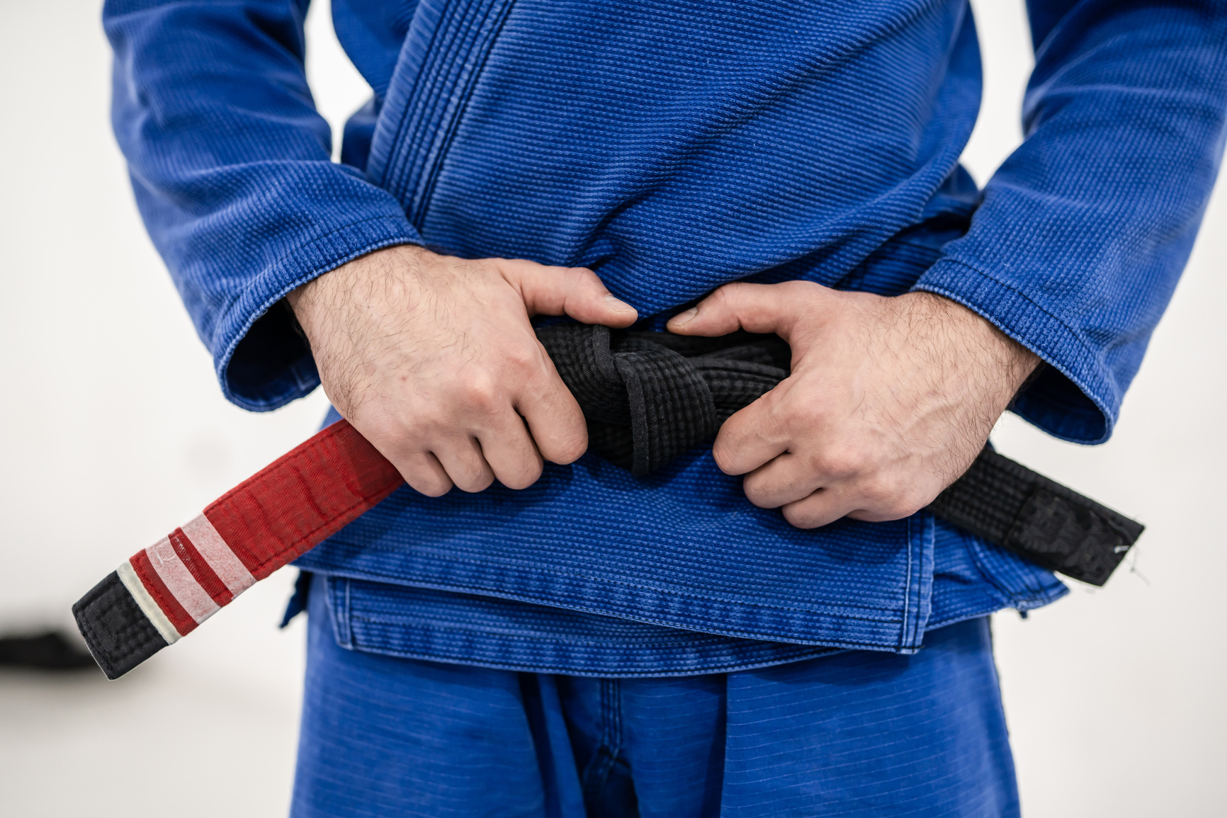 Close up on midsection of unknown caucasian male athlete bjj brazilian jiu-jitsu black belt standing on the mat at academy holding his belt during training wearing blue gi front view
