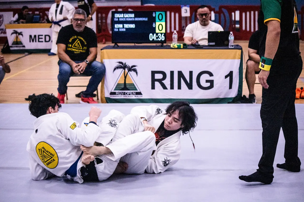 Two competitors in white gis grapple on the mat at Ring 1 while a referee watches and the scoreboard runs the clock.