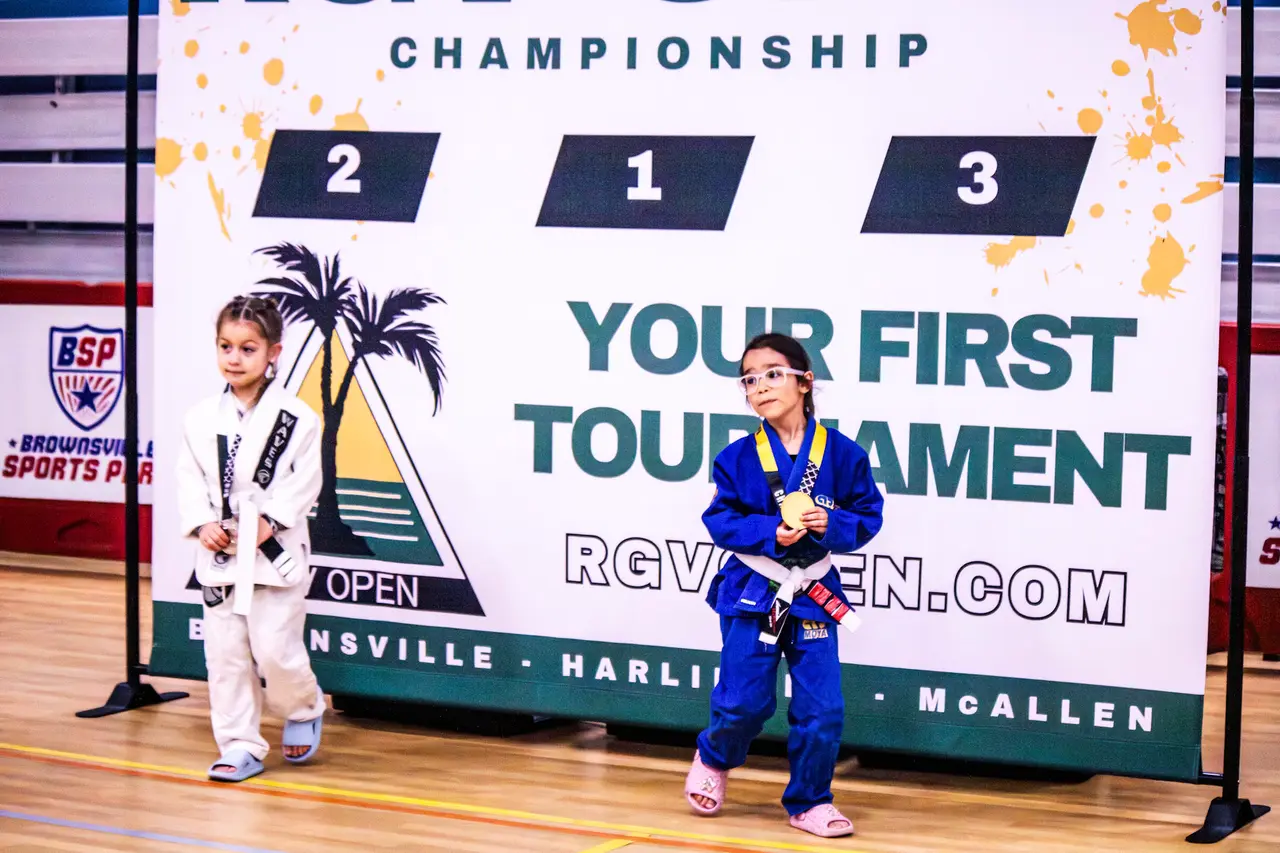 Two young competitors in gis with medals on the podium in front of the RGV Open “Your First Tournament” backdrop.